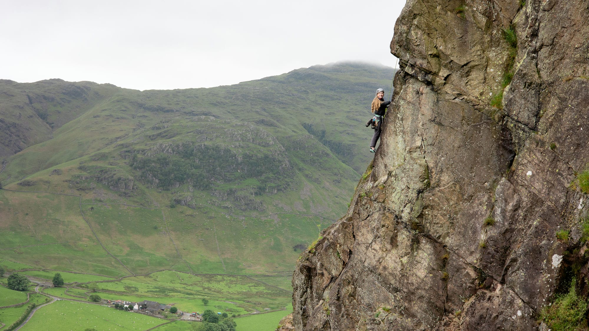a wide shot of someone half way up a guided rock climb showing the mountains in the background in the lake district