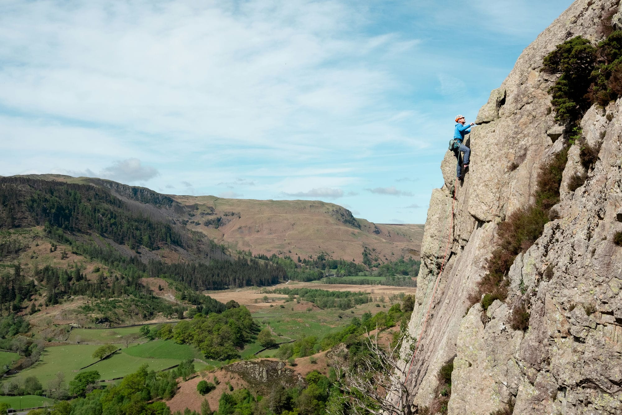 wide shot showing a climber at the top of a traditional rock climb with beautiful views of the lake district behind them